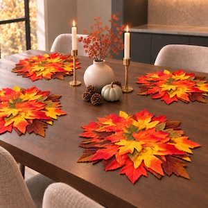 Peut inclure: Un ensemble de quatre sets de table ronds composés de feuilles d'automne artificielles superposées dans des tons rouge, orange, jaune et vert. Le centre de table comprend un vase en céramique blanche avec du feuillage orange et deux bougies.