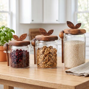May include: Three clear glass storage jars of varying sizes with wooden lids featuring a carved leaf design. Each jar includes a wooden spoon attached to the side. The jars are filled with dried cranberries, granola, and white rice respectively.