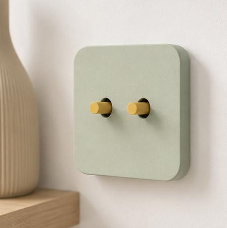 May include: A square light switch plate in a matte sage green finish featuring two cylindrical yellow toggle switches. The plate has rounded corners and a flat surface, providing a clean and functional interface for controlling electrical lighting.