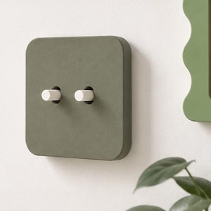 May include: A square, matte olive green light switch plate featuring two cylindrical white toggle switches. The plate has rounded corners and a smooth, flat surface, mounted flush against a plain, light-colored wall surface.