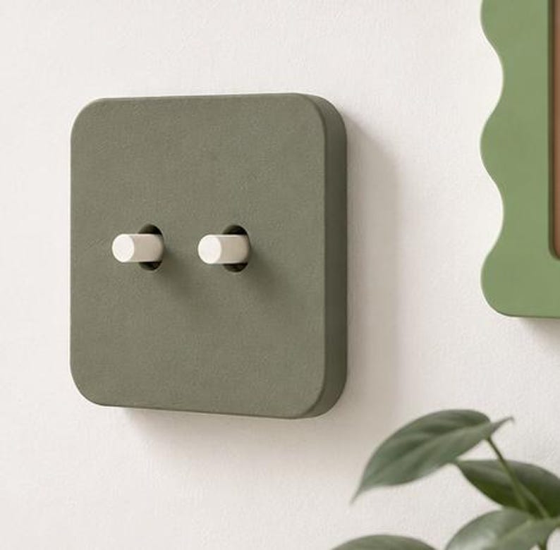 May include: A square, matte olive green light switch plate featuring two cylindrical white toggle switches. The plate has rounded corners and a smooth, flat surface, mounted flush against a plain, light-colored wall surface.