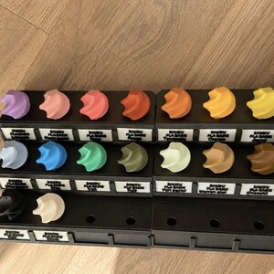 K&ouml;nnte beinhalten: Eine Sammlung von 3D-gedruckten Filament-Farbmustern, angeordnet in einem schwarzen Pr&auml;sentationsst&auml;nder. Jedes Muster hat eine einzigartige Farbe, darunter Violett, Rosa, Rot, Orange, Gelb, Blau, Gr&uuml;n, Braun, Schwarz und Wei&szlig;, mit Beschriftungen.