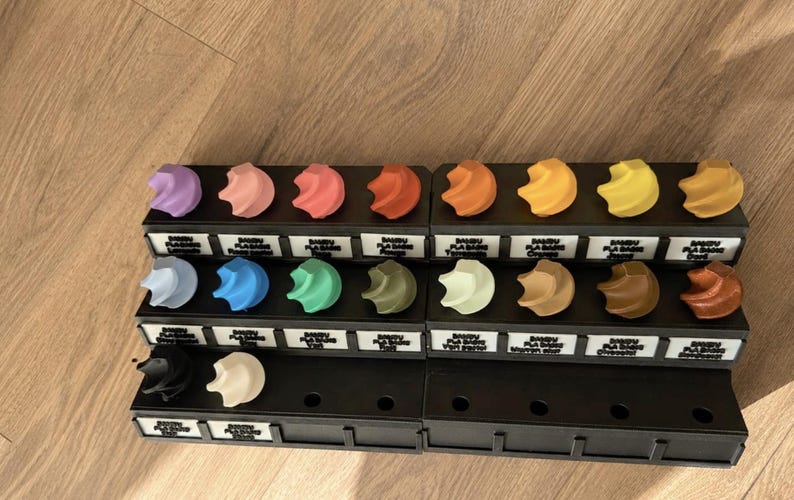 K&ouml;nnte beinhalten: Eine Sammlung von 3D-gedruckten Filament-Farbmustern, angeordnet in einem schwarzen Pr&auml;sentationsst&auml;nder. Jedes Muster hat eine einzigartige Farbe, darunter Violett, Rosa, Rot, Orange, Gelb, Blau, Gr&uuml;n, Braun, Schwarz und Wei&szlig;, mit Beschriftungen.