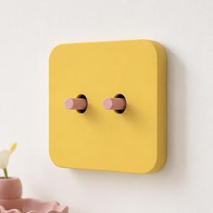 May include: A square yellow light switch plate with rounded corners featuring two cylindrical pink toggle switches protruding from the center. The switch plate is mounted on a flat surface and has a smooth, matte finish across the entire face.