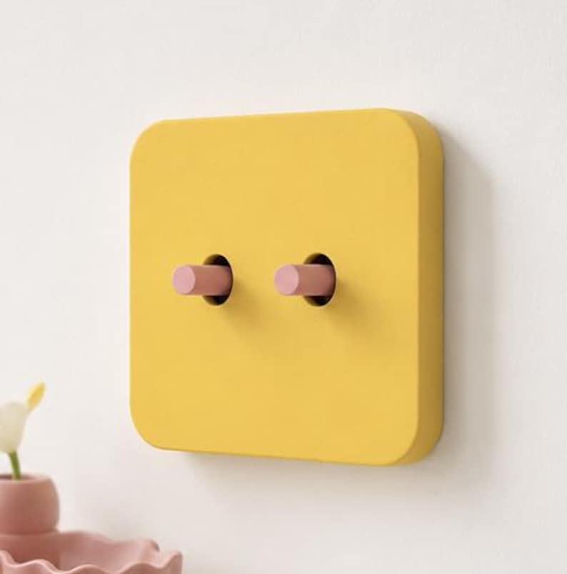 May include: A square yellow light switch plate with rounded corners featuring two cylindrical pink toggle switches protruding from the center. The switch plate is mounted on a flat surface and has a smooth, matte finish across the entire face.