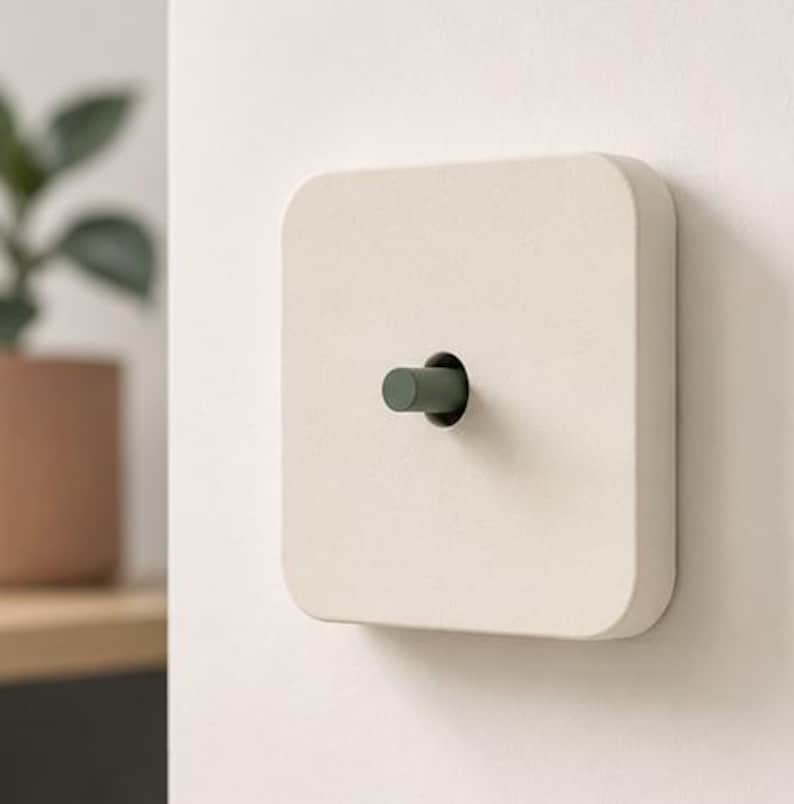 May include: A square light switch with a matte off-white finish and rounded corners. The switch features a central cylindrical toggle lever in a muted sage green color, protruding from a circular cutout in the center of the faceplate.