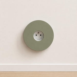 K&ouml;nnte beinhalten: Eine kreisf&ouml;rmige, salbeigr&uuml;ne Wandplatte mit einer mittig platzierten wei&szlig;en Steckdose. Die Platte hat eine glatte, matte Oberfl&auml;che und ist b&uuml;ndig an einer schlichten, hellen Wand montiert, was f&uuml;r eine saubere Optik beim Stromanschluss sorgt.