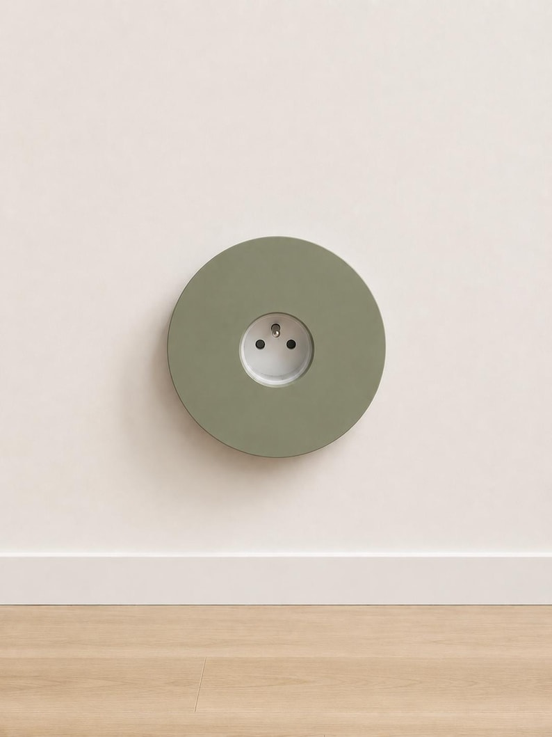 K&ouml;nnte beinhalten: Eine kreisf&ouml;rmige, salbeigr&uuml;ne Wandplatte mit einer mittig platzierten wei&szlig;en Steckdose. Die Platte hat eine glatte, matte Oberfl&auml;che und ist b&uuml;ndig an einer schlichten, hellen Wand montiert, was f&uuml;r eine saubere Optik beim Stromanschluss sorgt.