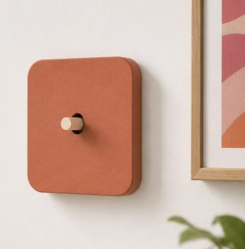 May include: A square light switch cover with a matte terracotta finish and rounded corners. The center features a cylindrical light-colored wooden toggle switch that protrudes from the face of the plate, mounted on a flat wall surface.