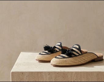 Puede incluir: Un par de mules sin talón confeccionadas en paja trenzada de tono beige natural, con rayas horizontales azul marino, punta redondeada y una borla negra decorativa en la parte superior de cada zapato, con un tacón bajo y plano marrón.