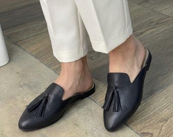 Puede incluir: Un par de mules de cuero negro con punta afilada y una borla decorativa en la parte superior de cada zapato. Los mules se llevan con pantalones blancos, mostrando un diseño sin talón, fácil de poner y con un perfil de tacón bajo.