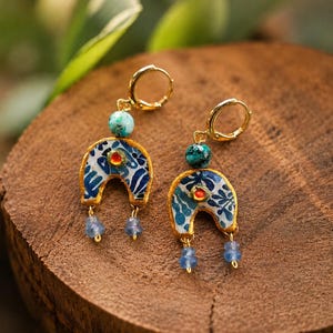 May include: A pair of gold-toned hoop earrings featuring a horseshoe-shaped pendant with a blue and white floral pattern. Each earring includes a turquoise-colored bead at the top, a small orange center accent, and two dangling light blue beads.