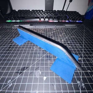May include: A blue fingerboard grind rail featuring a curved metal top bar supported by two blue rectangular bases. The structure is positioned on a black cutting mat with white grid lines, showcasing a functional design for finger skateboarding.