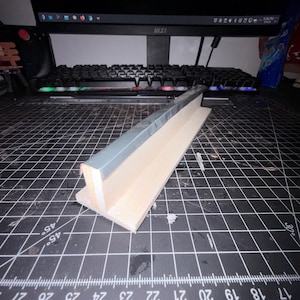May include: A fingerboard grind rail featuring a light-colored wooden base and a smooth, metallic grey top edge. The structure is T-shaped, designed for miniature skateboard tricks, and is positioned on a black cutting mat with white grid lines.