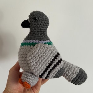 May include: A crocheted pigeon plush toy featuring a dark grey head, a light grey body, and a black beak. The bird has a green and light purple neck band, black wing stripes, and orange feet. The toy is crafted with visible yarn stitches.