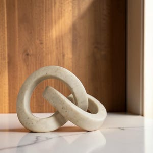 May include: A decorative sculpture consisting of two interlocking, rounded links made of a light beige, speckled stone material. The object features a smooth, polished surface with subtle grey and tan flecks throughout the solid, curved forms.