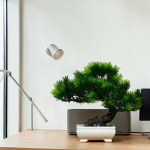 May include: A small artificial bonsai tree with vibrant green needle-like foliage and a dark textured trunk. The tree is planted in a rectangular white ceramic pot with four small feet, positioned on a light-coloured wooden surface.