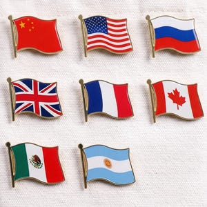 Peut inclure: Un ensemble de huit épinglettes en émail métallique en forme de drapeaux flottants, incluant ceux de la Chine, des États-Unis, de la Russie, du Royaume-Uni, de la France, du Canada, du Mexique et de l'Argentine, fixés sur un tissu blanc cassé.