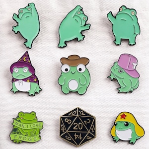 May include: A collection of nine enamel pins featuring various green frogs in different poses and outfits, including a wizard, a cowboy, and a baseball cap wearer, alongside a black twenty-sided die pin and a frog with a banner reading MAN, I LOVE FROGS.