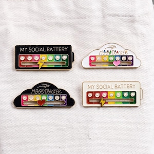 Puede incluir: Cuatro pines de esmalte con indicadores deslizantes. Dos pines rectangulares dicen MY SOCIAL BATTERY con un rayo amarillo. Dos pines en forma de nube dicen MY MOODTRACKER con un corazón rosa o rojo. Cada uno tiene una escala de arcoíris.