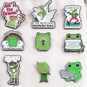 May include: A collection of nine enamel pins featuring green frogs in various poses and outfits. The pins include text such as Am I the Drama, I am up to some NONSENSE, STRAWBERRY JAMS BUT MY GLOCK DON'T, breathe...relax exhale the fuckery, Not Now Partner My Tummy Hurts, NEVER FROGET I LOVE YOU, and JUST DOING MY SILLY LITTLE TASKS.