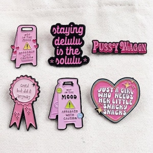 May include: Six enamel pins in shades of pink, black, and yellow. Designs include two caution signs reading "NOT IN THE MOOD APPROACH WITH CAUTION," a ribbon badge saying "Cried but did it anyway," text reading "staying delulu is the solulu," "Pussy Wagon," and a heart saying "JUST A GIRL WHO NEEDS HER LITTLE SNACKY SNACKS."