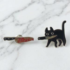 Peut inclure: Deux broches en émail représentant un chat noir tenant un couteau dans sa gueule et une broche en forme de couteau avec le texte rouge "Killing it" écrit en écriture cursive. Les deux broches ont une finition noire brillante et des contours argentés.