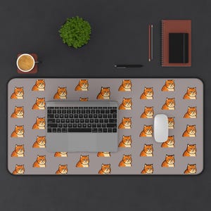May include: A rectangular desk mat featuring a repeating pattern of grumpy orange tabby cat faces on a light grey background. The mat is positioned under a silver laptop and a white computer mouse, showcasing a consistent cartoon cat design.