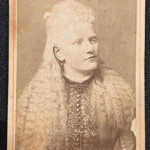 May include: A sepia-toned vintage cabinet card photograph featuring a portrait of a person with long, wavy, light-colored hair styled half-up. The individual wears a high-collared, dark-colored garment with a vertical row of buttons. The text Osenmann New York is printed at the bottom.