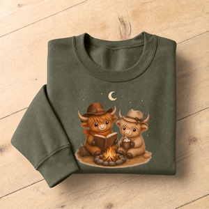 May include: An olive green crewneck sweatshirt featuring a graphic of two cartoon highland cows wearing cowboy hats sitting by a campfire at night. One cow reads an open book while the other holds a mug with marshmallows under a crescent moon.