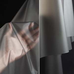 Może przedstawiać: Półprzezroczysty, półmatowy arkusz elastycznego materiału o gładkim wykończeniu. Materiał jest cienki i giętki, przepuszcza światło, jednocześnie zasłaniając znajdujące się za nim detale, tworząc miękkie fałdy oraz delikatne, płynne krzywizny.