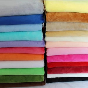 May include: A collection of folded fabric squares stacked in two vertical columns. The fabrics feature a soft, plush texture and a wide variety of solid colours, including shades of grey, blue, teal, purple, pink, orange, yellow, brown, red, green, and black.