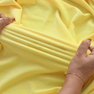 May include: A solid, bright yellow fabric with a smooth, matte texture. The material is being stretched horizontally, creating deep, parallel folds and ripples across the center of the textile, demonstrating its flexible and elastic nature.