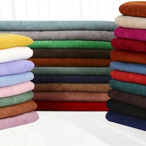 May include: A collection of folded corduroy fabric pieces stacked in three separate piles. The fabric features a distinct vertical ribbed texture and comes in a wide variety of solid colours including yellow, white, blue, pink, green, brown, red, black, and grey.