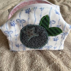 May include: A fabric pouch featuring a white background with a blue ribbon and floral pattern. The front displays a dark gray circular applique with a star-shaped center and two green striped leaves. The pouch has a white zipper and pink lining.