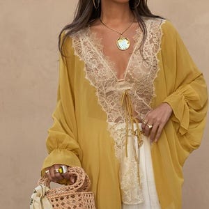 May include: A mustard yellow sheer blouse featuring long, loose sleeves with ruffled cuffs and a deep V-neckline trimmed with intricate beige lace. The front includes a delicate lace panel and thin tie closures. Paired with white trousers and a woven bag.