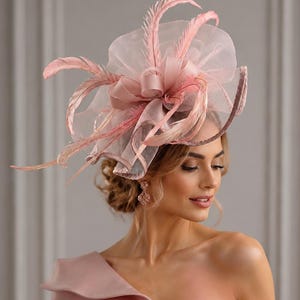 May include: A dusty pink fascinator hat featuring layered sheer organza loops, delicate upright feathers, and a structured base. The headpiece is paired with a matching dusty pink one-shoulder dress with a folded neckline detail and subtle pleating.