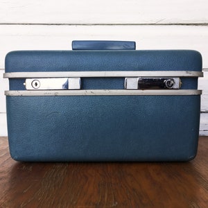 Könnte beinhalten: Ein vintage blauer Hartschalenkoffer mit silbernen Akzenten und zwei Schlössern. Der Koffer hat eine strukturierte Oberfläche und einen Griff oben.