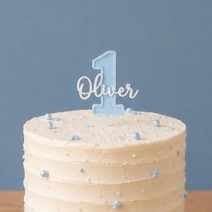 May include: A round white frosted cake decorated with small light blue edible pearls and star-shaped sprinkles. A light blue cake topper shaped like the number one with the name Oliver in white script lettering is inserted into the top.
