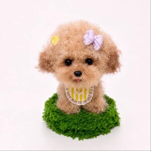 May include: A tan, fluffy, needle-felted toy poodle figurine with black eyes and a black nose. It wears a yellow and white striped bib with a white lace trim, a small yellow hair clip, and a light purple bow hair accessory, sitting on a green base.