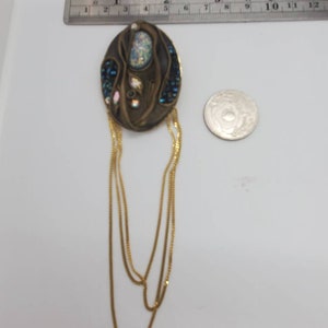 May include: An oval-shaped, antique-style brooch with a bronze finish. The brooch features an iridescent central stone, surrounded by blue and iridescent crystals. Gold chains hang from the bottom. A ruler shows the brooch is approximately 2 inches long.