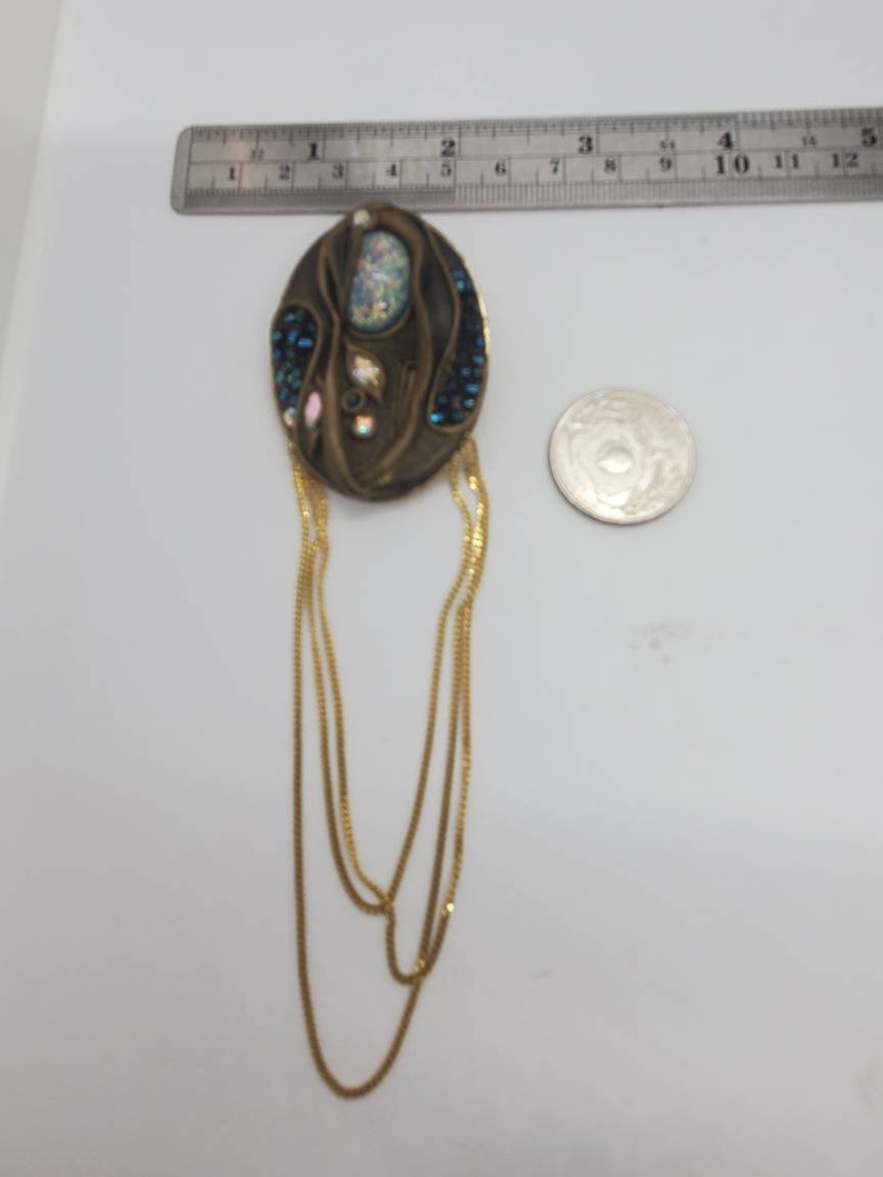 May include: An oval-shaped, antique-style brooch with a bronze finish. The brooch features an iridescent central stone, surrounded by blue and iridescent crystals. Gold chains hang from the bottom. A ruler shows the brooch is approximately 2 inches long.