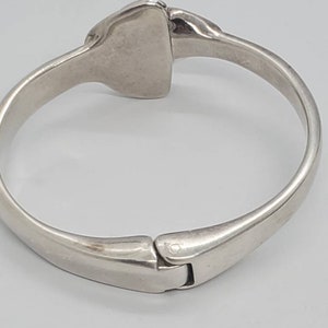 May include: A silver-toned hinged bracelet with a rectangular centerpiece. The bracelet has a smooth, polished finish and a clasp closure. The design is simple and elegant, suitable for everyday wear.