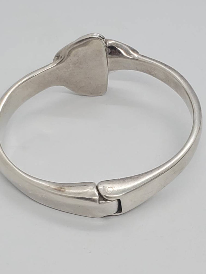 May include: A silver-toned hinged bracelet with a rectangular centerpiece. The bracelet has a smooth, polished finish and a clasp closure. The design is simple and elegant, suitable for everyday wear.