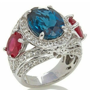 May include: A silver ring featuring a large, oval, teal-colored gemstone as the centerpiece, flanked by two smaller, red gemstones. The ring is encrusted with numerous small, clear gemstones, adding sparkle and detail to the design.