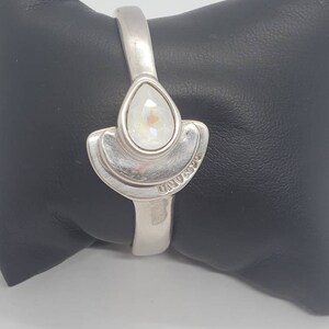 May include: A silver-toned hinged bracelet with a pear-shaped, iridescent crystal centerpiece. The crystal is set within a stylized, fan-shaped design. The word "UNOAERRE" is engraved on the bracelet. The bracelet is displayed on a black surface.