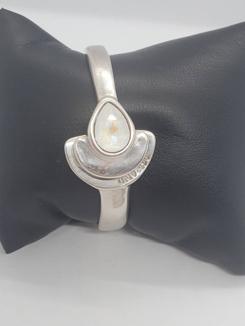 May include: A silver-toned hinged bracelet with a pear-shaped, iridescent crystal centerpiece. The crystal is set within a stylized, fan-shaped design. The word "UNOAERRE" is engraved on the bracelet. The bracelet is displayed on a black surface.