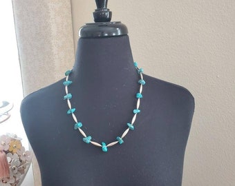Puede incluir: Un collar de piedras turquesas expuesto sobre un maniquí negro. El collar presenta piedras irregulares de color azul verdoso brillante intercaladas con pequeñas cuentas cilíndricas de color plateado. El collar es de diseño sencillo y elegante.