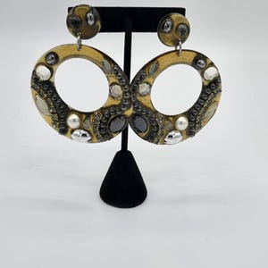 May include: A pair of large, oval-shaped earrings with a gold-toned base. The earrings are adorned with various embellishments, including clear, black, and silver-colored stones, and small pearls. The earrings are displayed on a black jewelry stand against a white background.