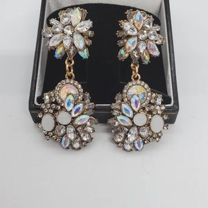 May include: A pair of gold-toned, dangling earrings with a floral design. Each earring features a cluster of clear and iridescent crystals, with a central white oval stone. The earrings are displayed in a black jewelry box.